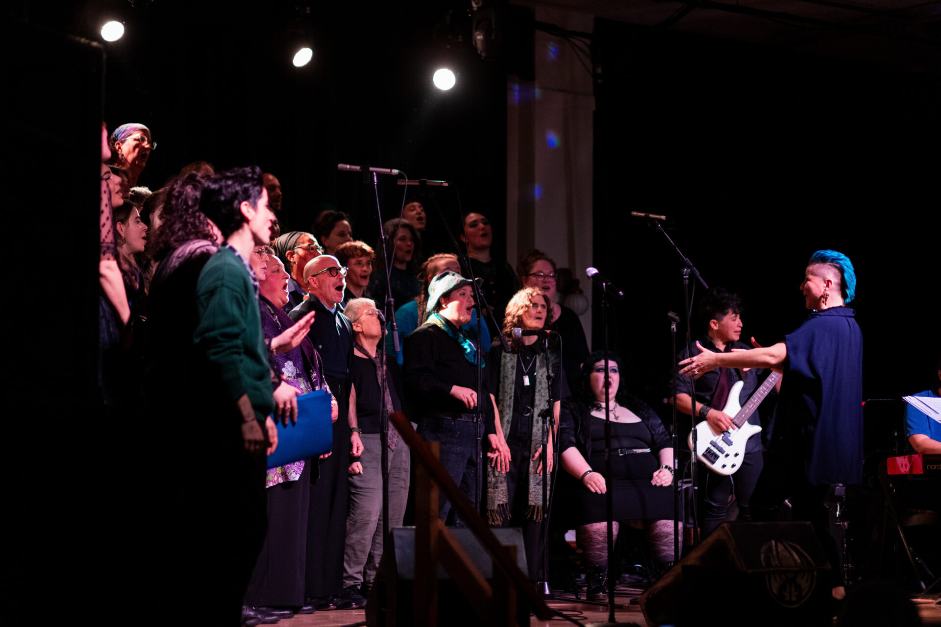 Image of elias conducting Queer Choir, an intergenerational group of queers standing on a stage wearing colors resembling those found on a peacock. There is joy and energy in everyone's faces!